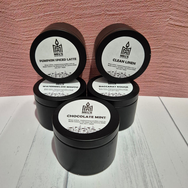 Five matte black candle tins with different labels, placed on a wooden surface against a pink background. The tins are closed and the labels face the front, showcasing various scents like Pumpkin Spice Latte, Clean Linen, Watermelon Seeds, and Chocolate Mint.
